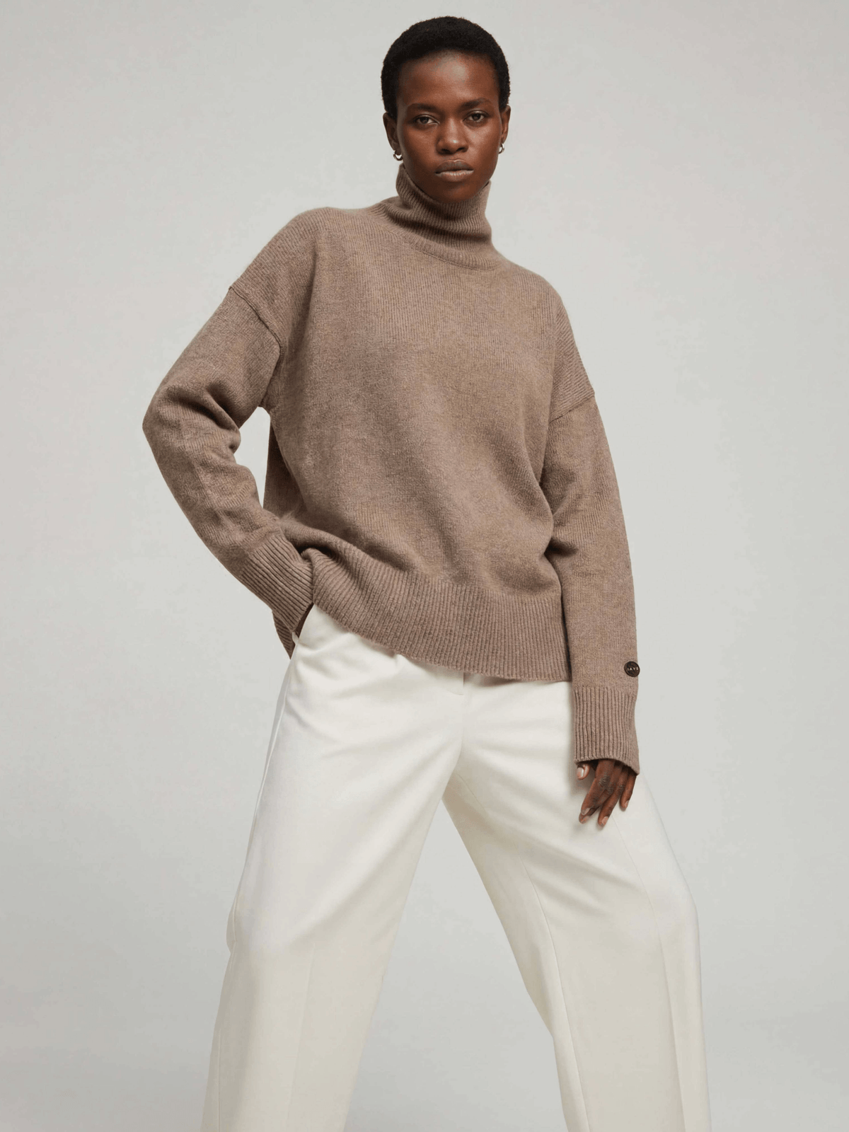 Woman wearing a brown sweater and white pants on a plain background